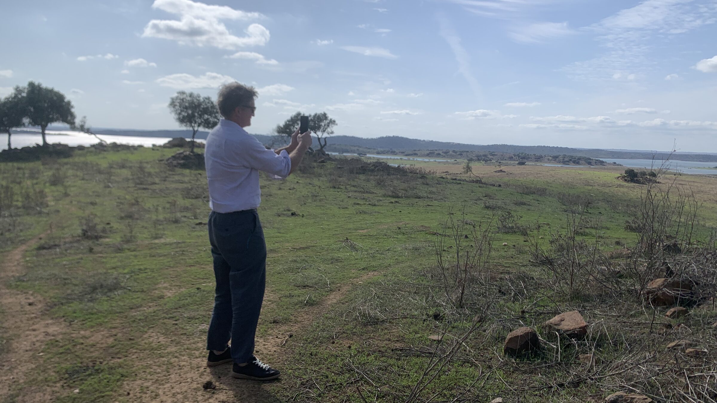 A figure photographing an open pastoral landscape with water in the distance.
