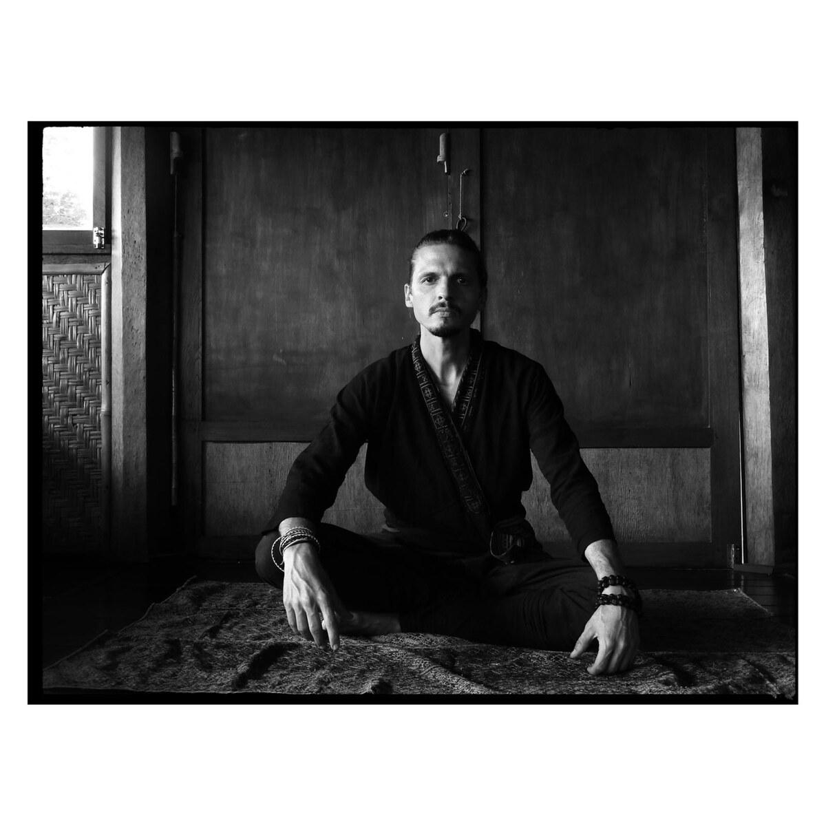 Black-and-white portrait of the practitioner seated cross-legged on a woven mat, looking directly at camera.